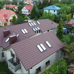 MASHOUSE Firma Budowlana - Nowy brązowy dach z blachodachówki, z licznymi oknami dachowymi i kominami, na domu o jasnych ścianach. W tle zielone otoczenie, obok stos starych materiałów dachowych.