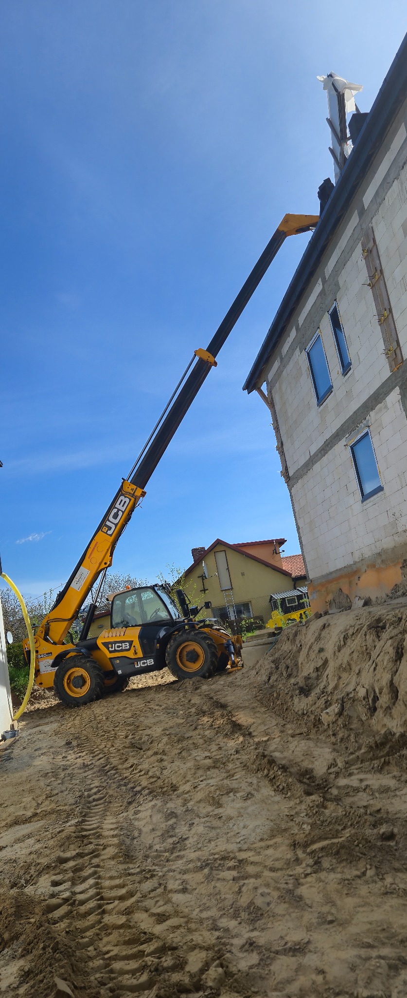 Żółty podnośnik teleskopowy JCB przy budynku z pustaków, prawdopodobnie podczas prac dekarskich. Widoczne okna i fragment dachu. Błękitne niebo w tle.