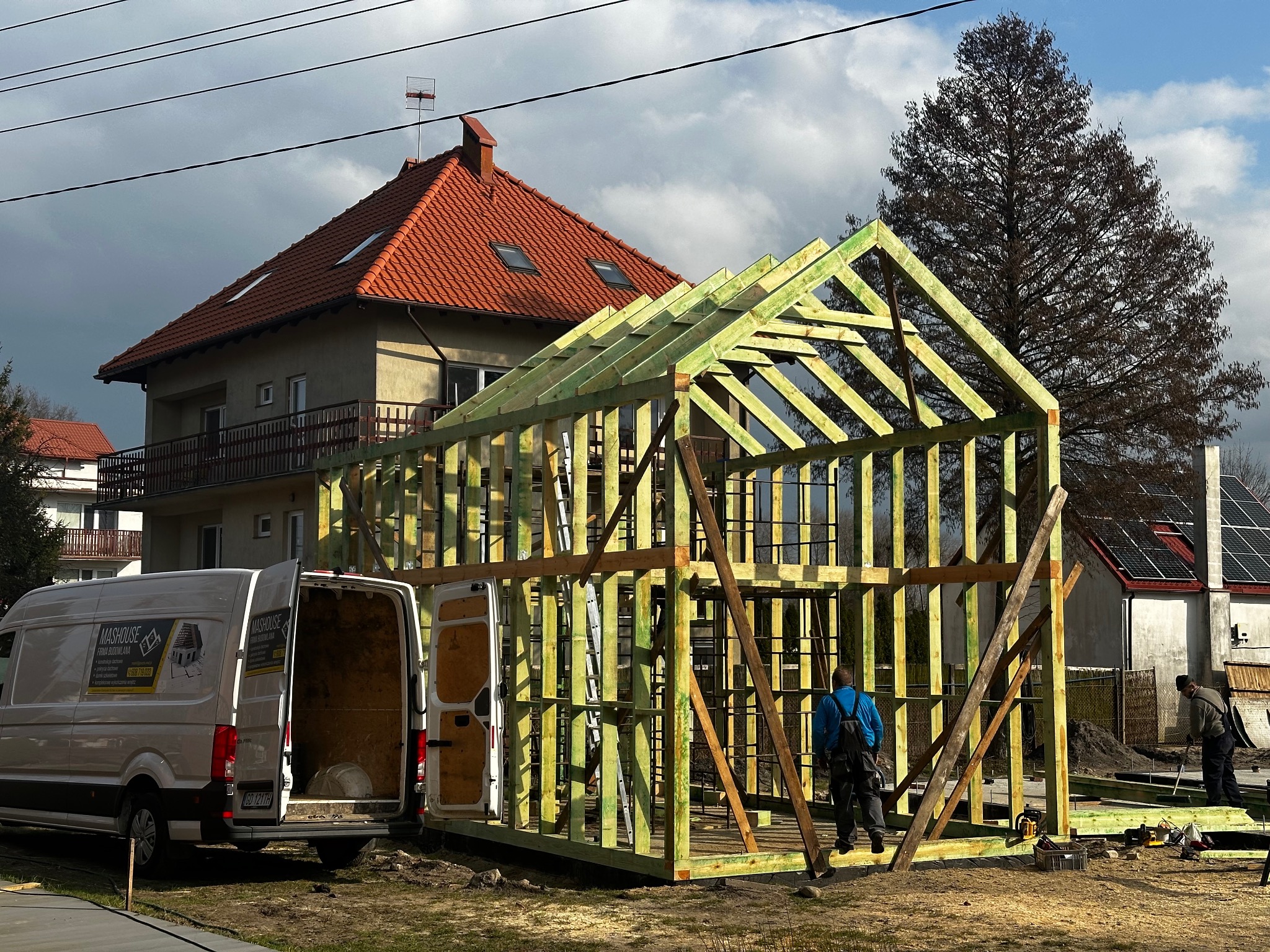 Wznoszenie szkieletu domu z drewna na tle budynku mieszkalnego z czerwoną dachówką. Na pierwszym planie biały bus z otwartymi drzwiami.