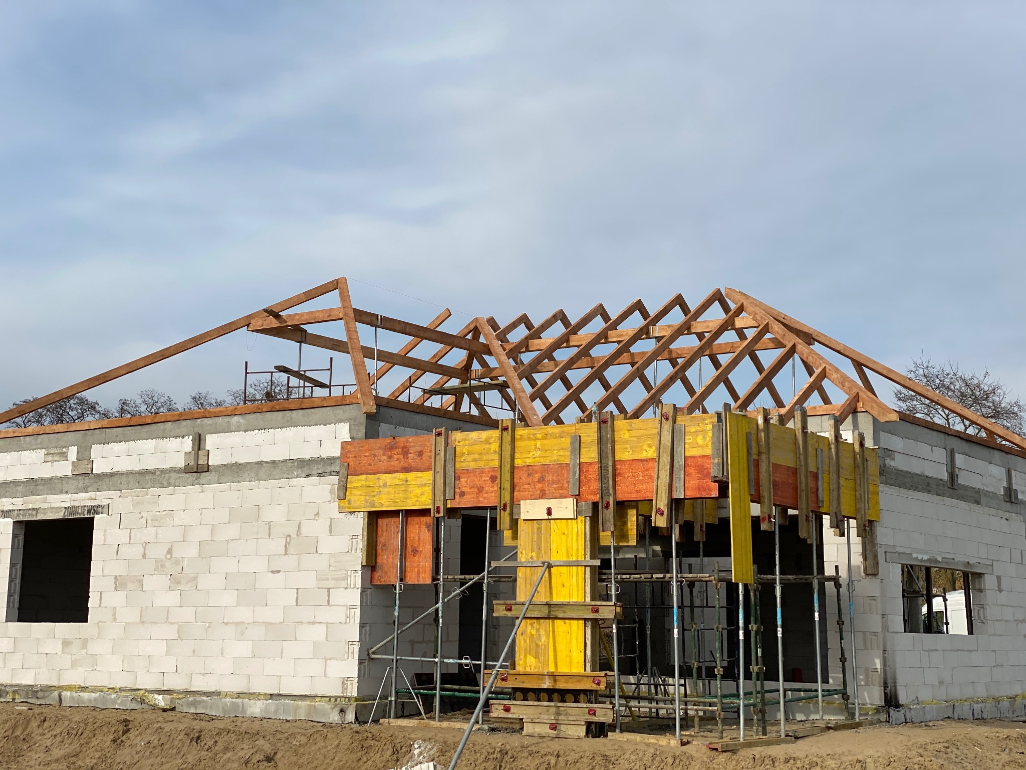 Budowa domu z białych bloczków: drewniana więźba dachowa, żółto-pomarańczowe szalunki okienne, rusztowania. Widok na wznoszony dom w stanie surowym otwartym.