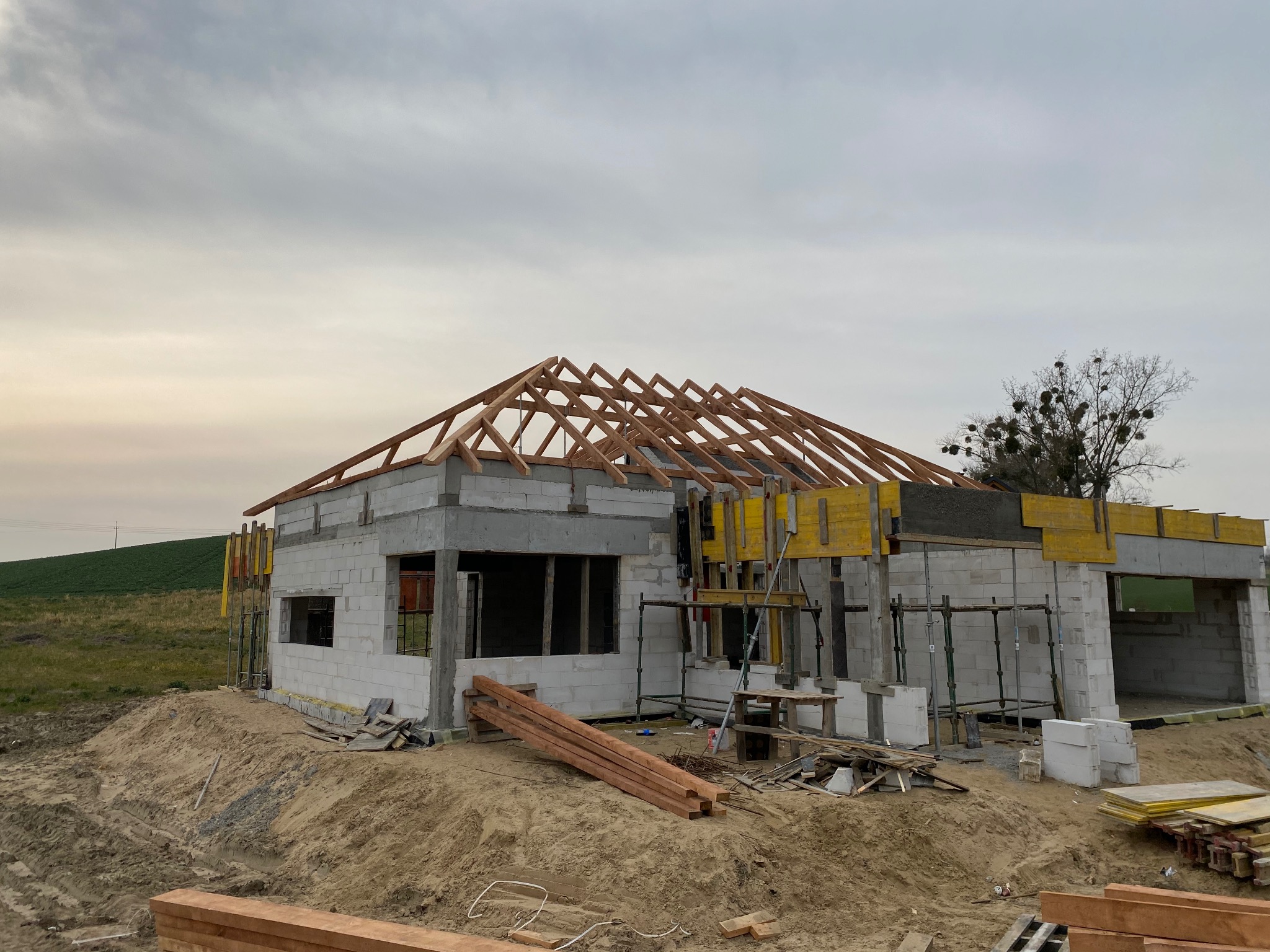 Budowa domu z białych bloczków: wzniesione ściany, drewniana więźba dachowa w trakcie montażu, szalunki i materiały budowlane na placu budowy.