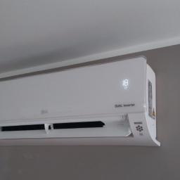 Sandblasting Mateusz Serwinek - Biała jednostka wewnętrzna klimatyzatora LG Dual Inverter zamontowana na szarej ścianie, wyświetlająca temperaturę 18 stopni Celsjusza, widoczna również kamera monitoringu pod sufitem.