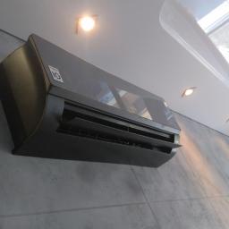 Sandblasting Mateusz Serwinek - Czarny klimatyzator LG zamontowany na szarej ścianie we Władysławowie, widoczny z dołu, z widocznym oświetleniem sufitowym i fragmentem okna.