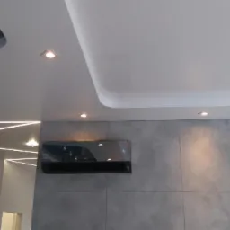 Nowoczesny klimatyzator ścienny w szarej obudowie, zamontowany na ścianie wykończonej płytami imitującymi beton, z widocznym oświetleniem sufitowym LED.