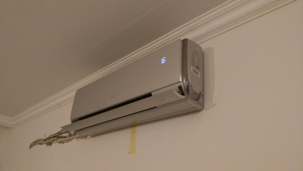 Szary klimatyzator ścienny z wyświetlaczem temperatury 16 stopni, zamontowany na ścianie z widocznymi śladami po instalacji.