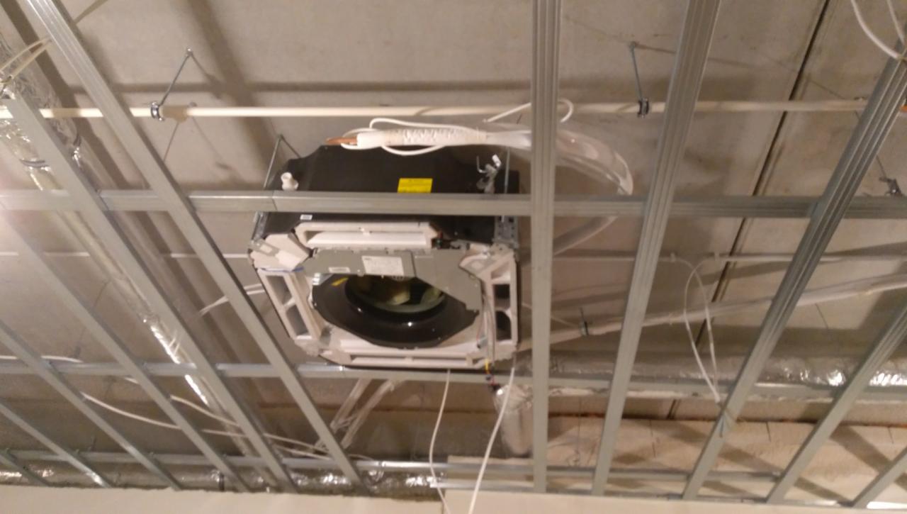 Kasetonowa jednostka wewnętrzna systemu klimatyzacji zamontowana w suficie podwieszanym z widocznymi przewodami i elementami konstrukcyjnymi.