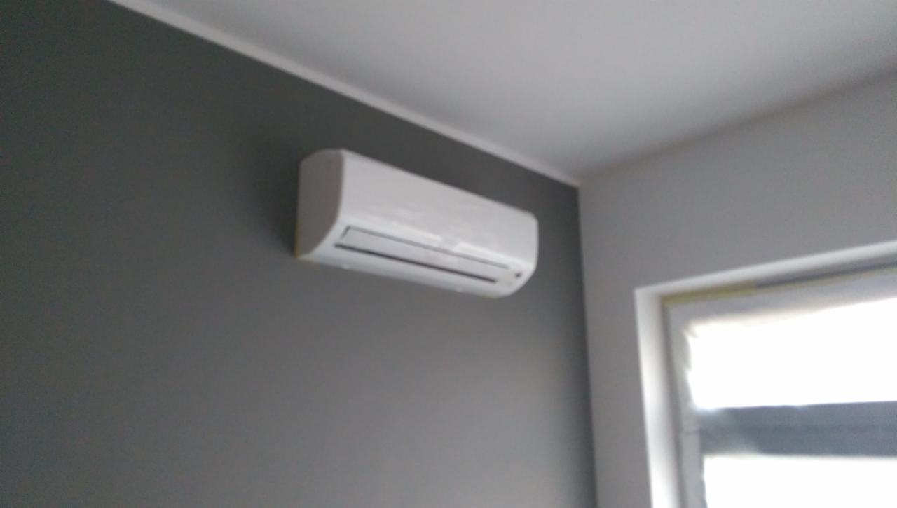 Biała jednostka klimatyzacji ściennej na szarej ścianie w pomieszczeniu z oknem w tle.