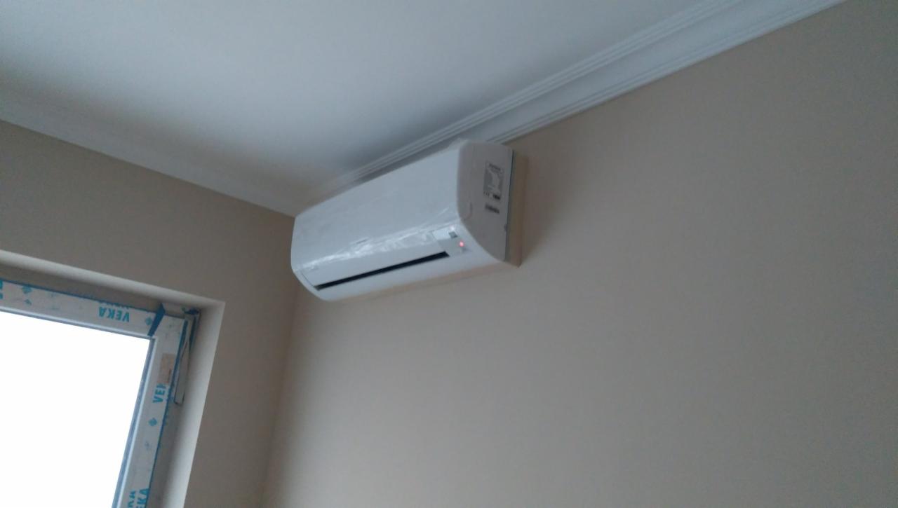 Świeżo zamontowana, biała jednostka wewnętrzna klimatyzatora na ścianie, jeszcze w folii ochronnej, widoczna rama okna z PCV marki Veka.