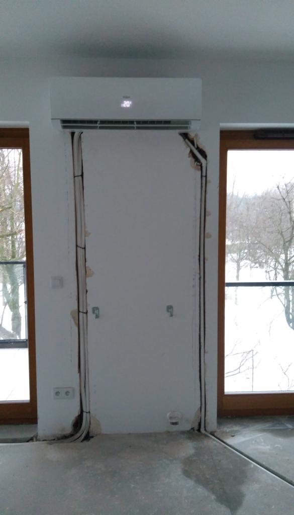 Biały klimatyzator ścienny z wyświetlaczem temperatury 20 stopni Celsjusza, zamontowany nad otworem drzwiowym w trakcie instalacji, widoczne kable i niedokończone wykończenie ściany, okna...