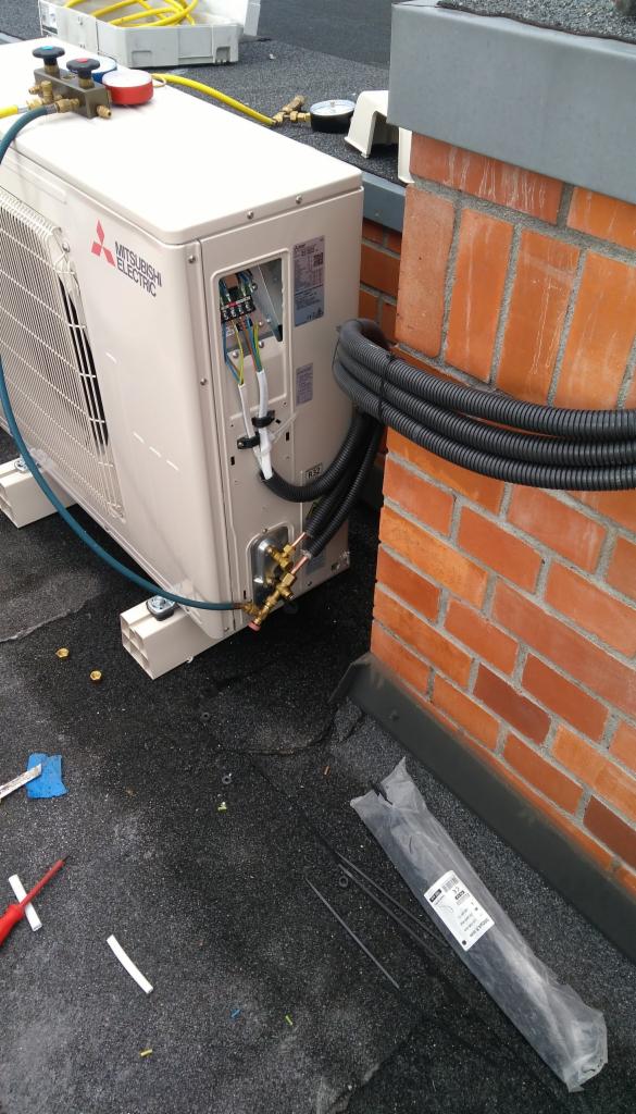 Jednostka zewnętrzna klimatyzacji Mitsubishi Electric zamontowana na dachu, z widocznymi przewodami instalacyjnymi i narzędziami w tle.