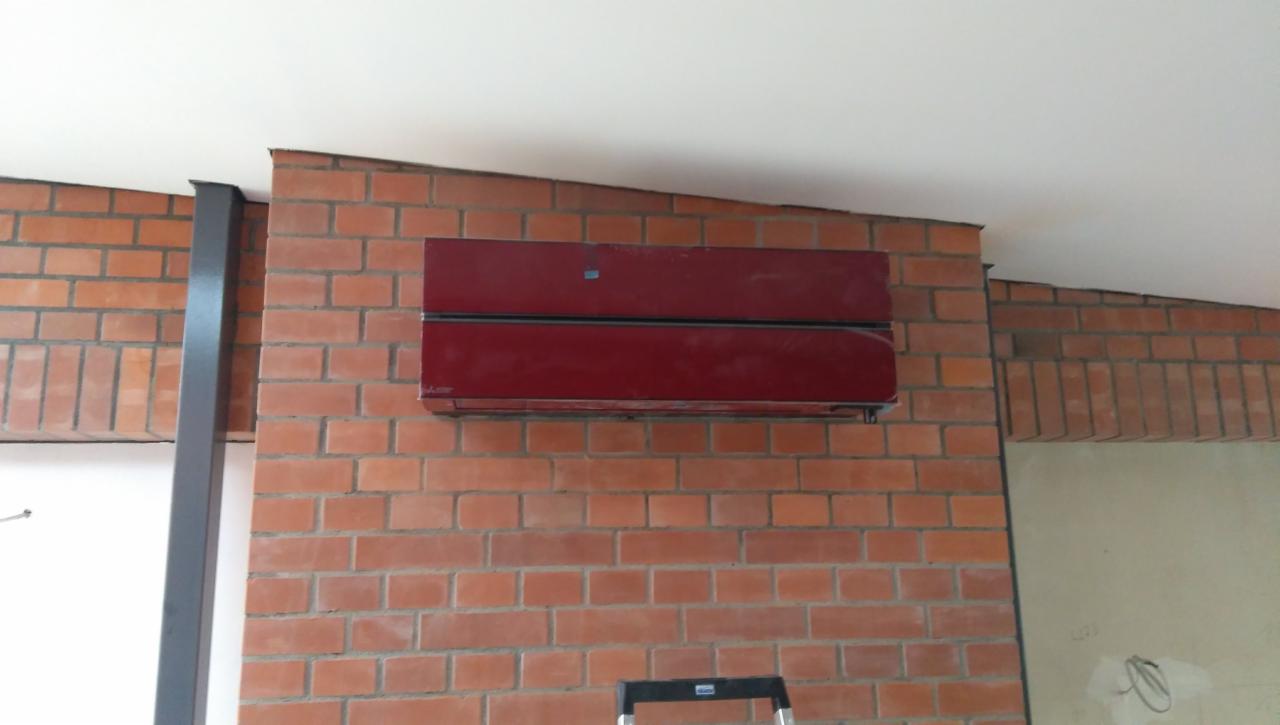 Czerwony klimatyzator Mitsubishi zamontowany na ścianie z cegły, z widoczną częścią drabiny w dolnej części kadru.