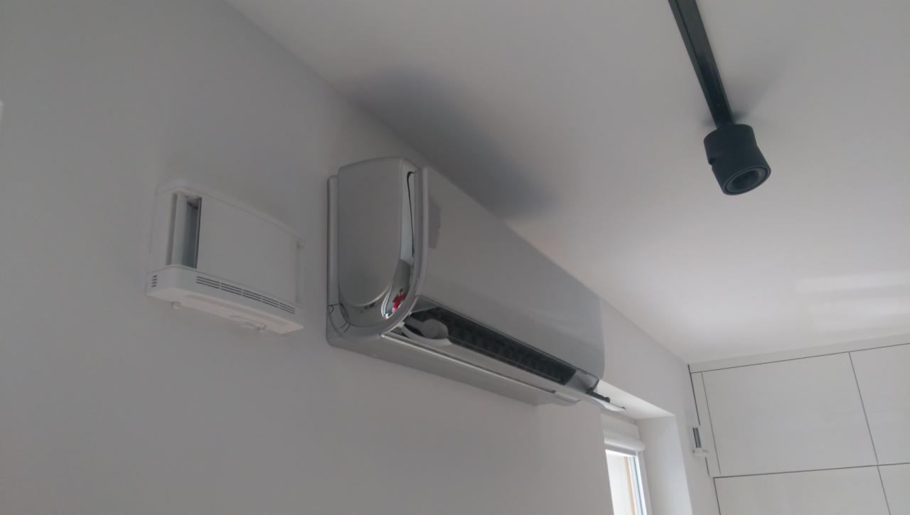 Nowoczesny klimatyzator ścienny z chromowanymi elementami, zamontowany w jasnym pomieszczeniu z oświetleniem punktowym na suficie i kratką wentylacyjną obok.