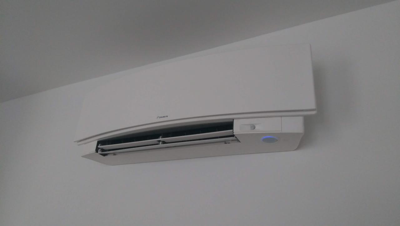 Biała klimatyzacja Daikin zamontowana na jasnej ścianie, widoczne żaluzje i niebieska dioda sygnalizacyjna.