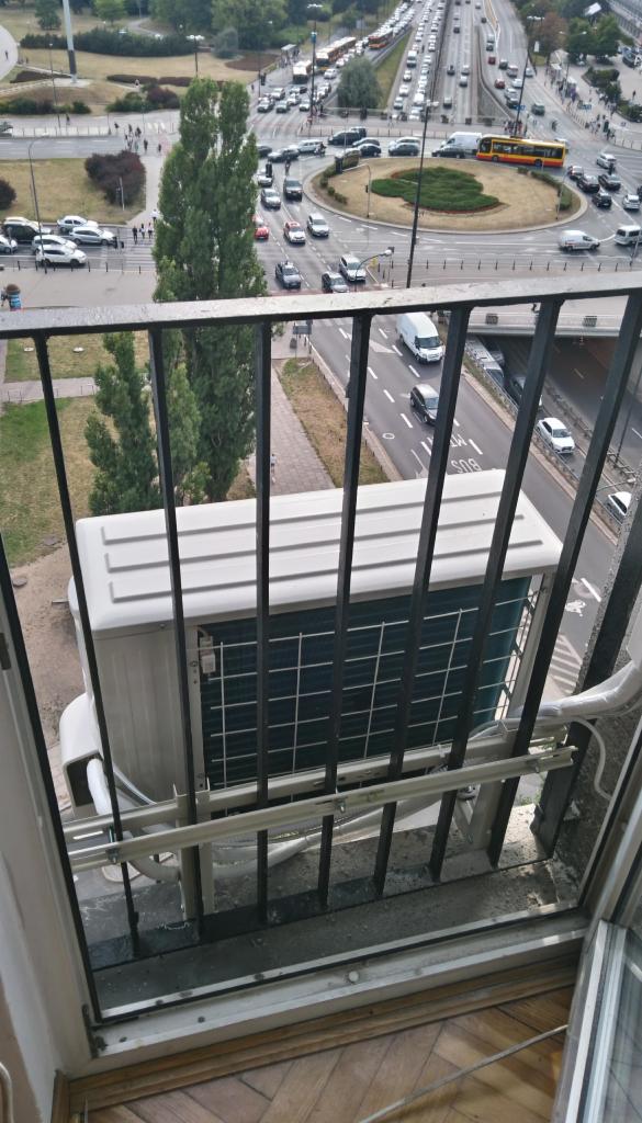 Zewnętrzna jednostka klimatyzatora zamontowana na balkonie z widokiem na ruchliwe rondo z perspektywy wysokiego piętra.