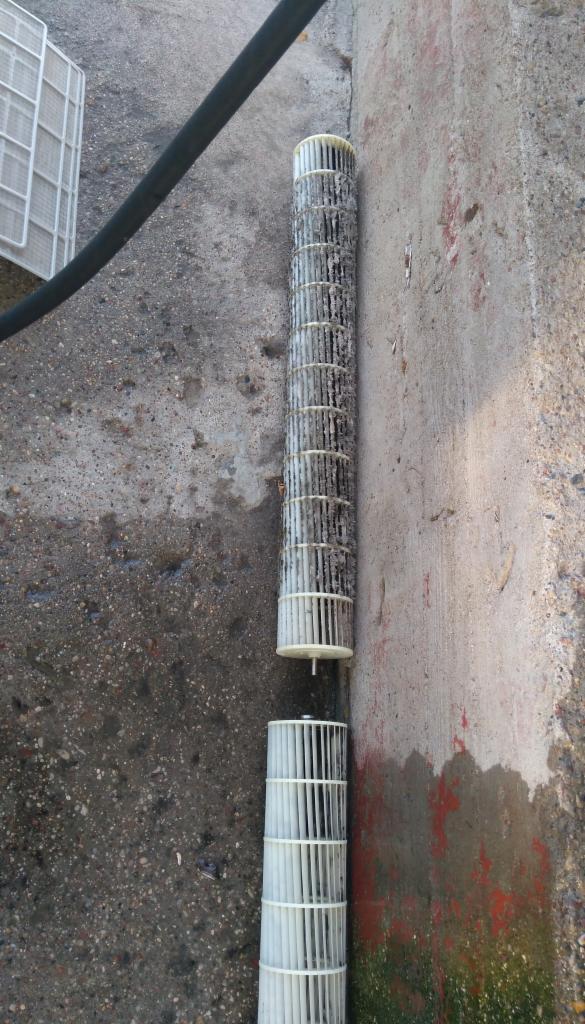 Dwa cylindryczne filtry wentylacyjne leżące na betonowym podłożu, jeden bardzo zabrudzony, drugi czysty, obok ściany z zaciekami.