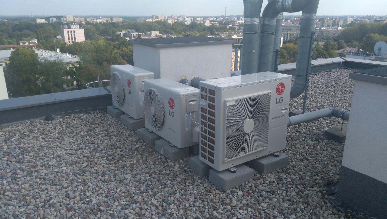 Trzy jednostki zewnętrzne LG, prawdopodobnie klimatyzacji, zamontowane na betonowych podstawach na dachu pokrytym żwirem, z widokiem na panoramę miasta w tle.