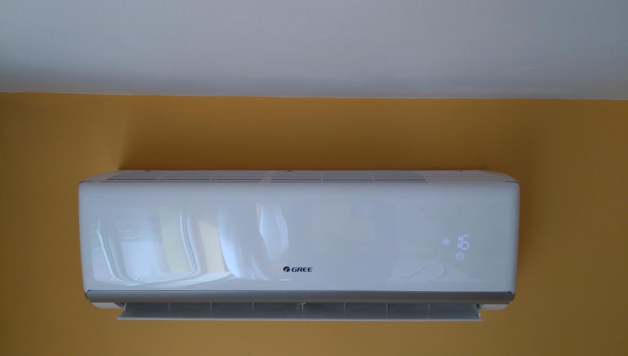 Biała jednostka wewnętrzna klimatyzatora marki Gree zamontowana na pomarańczowej ścianie, wyświetlająca temperaturę 16 stopni Celsjusza.