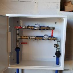 KRET-INSTAL Waldemar Szczepański - Wewnętrzny widok białej skrzynki z dwoma licznikami wody, zaworami kulowymi z czerwonymi uchwytami, rurami z niebieskiego tworzywa sztucznego i metalowymi złączkami, zamontowanej na surowej ścianie...