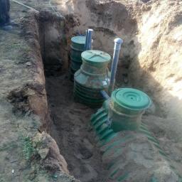 KRET-INSTAL Waldemar Szczepański - Montaż przydomowej oczyszczalni ścieków: zielone zbiorniki z pokrywami w wykopie, częściowo zasypane piaskiem, widoczne rury przyłączeniowe.