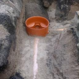 KRET-INSTAL Waldemar Szczepański - Pomarańczowa studzienka rewizyjna umieszczona w wykopie o nierównych ścianach, widoczny ślad po rurze na dnie wykopu.