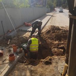 KRET-INSTAL Waldemar Szczepański - Dwóch robotników pracuje w wykopie przy krawężniku, jeden kopie łopatą, drugi stoi w żółtej kamizelce odblaskowej, widoczne rury PCV, w tle billboard i ruch uliczny.