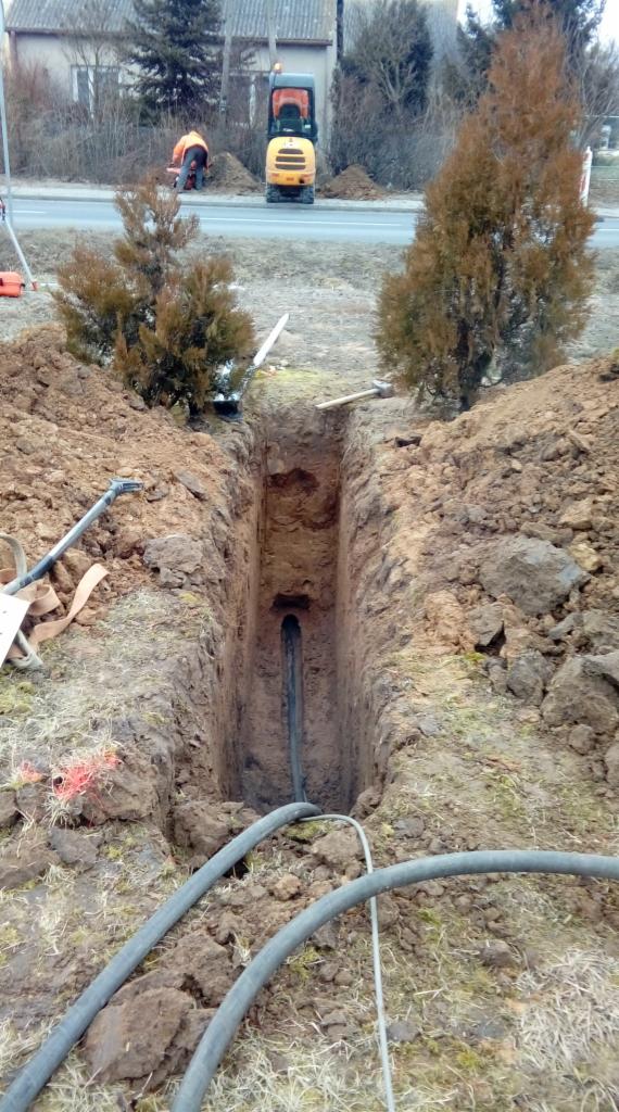 Wykopany rów z widoczną rurą na dnie, na zewnątrz rury leżą kable, w tle mini koparka i pracownik w pomarańczowej kamizelce.