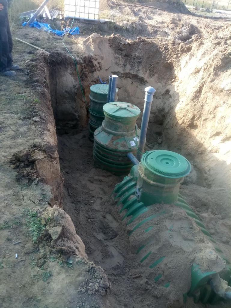 Montaż przydomowej oczyszczalni ścieków: zielone zbiorniki z pokrywami w wykopie, częściowo zasypane piaskiem, widoczne rury przyłączeniowe.