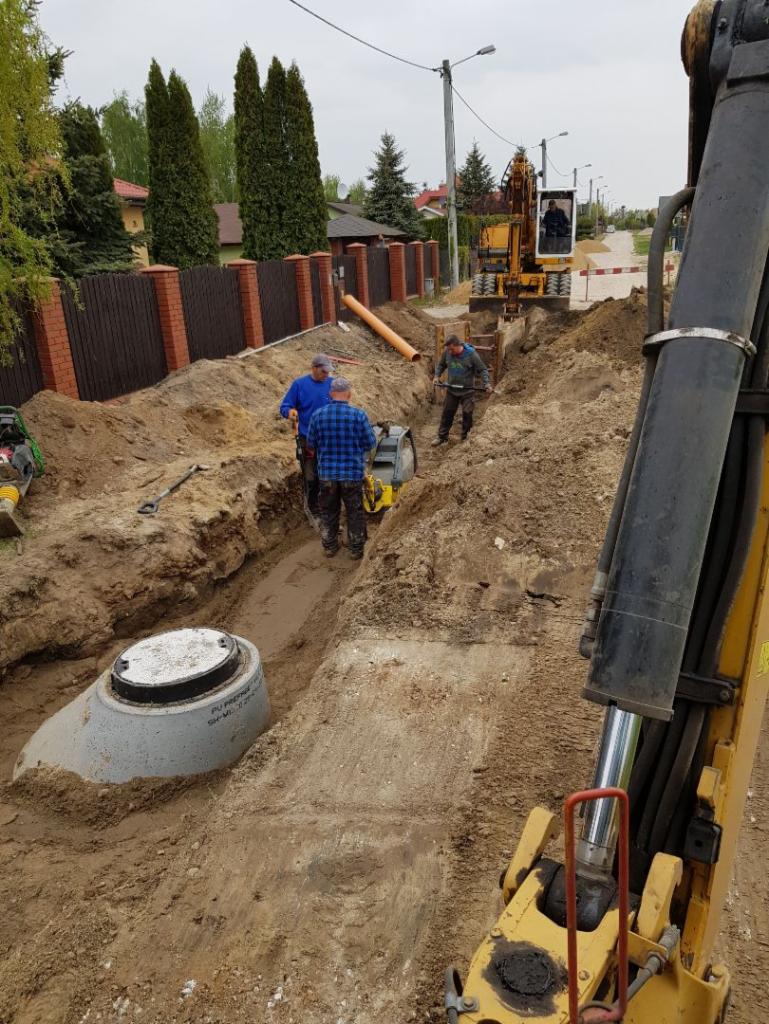 Betonowy zbiornik w wykopie, obok pracownicy z ubijarką i koparka w tle, rury kanalizacyjne na powierzchni, etap prac ziemnych.