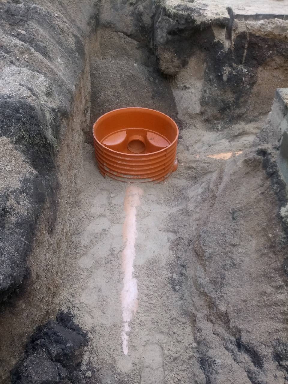 Pomarańczowa studzienka rewizyjna umieszczona w wykopie o nierównych ścianach, widoczny ślad po rurze na dnie wykopu.