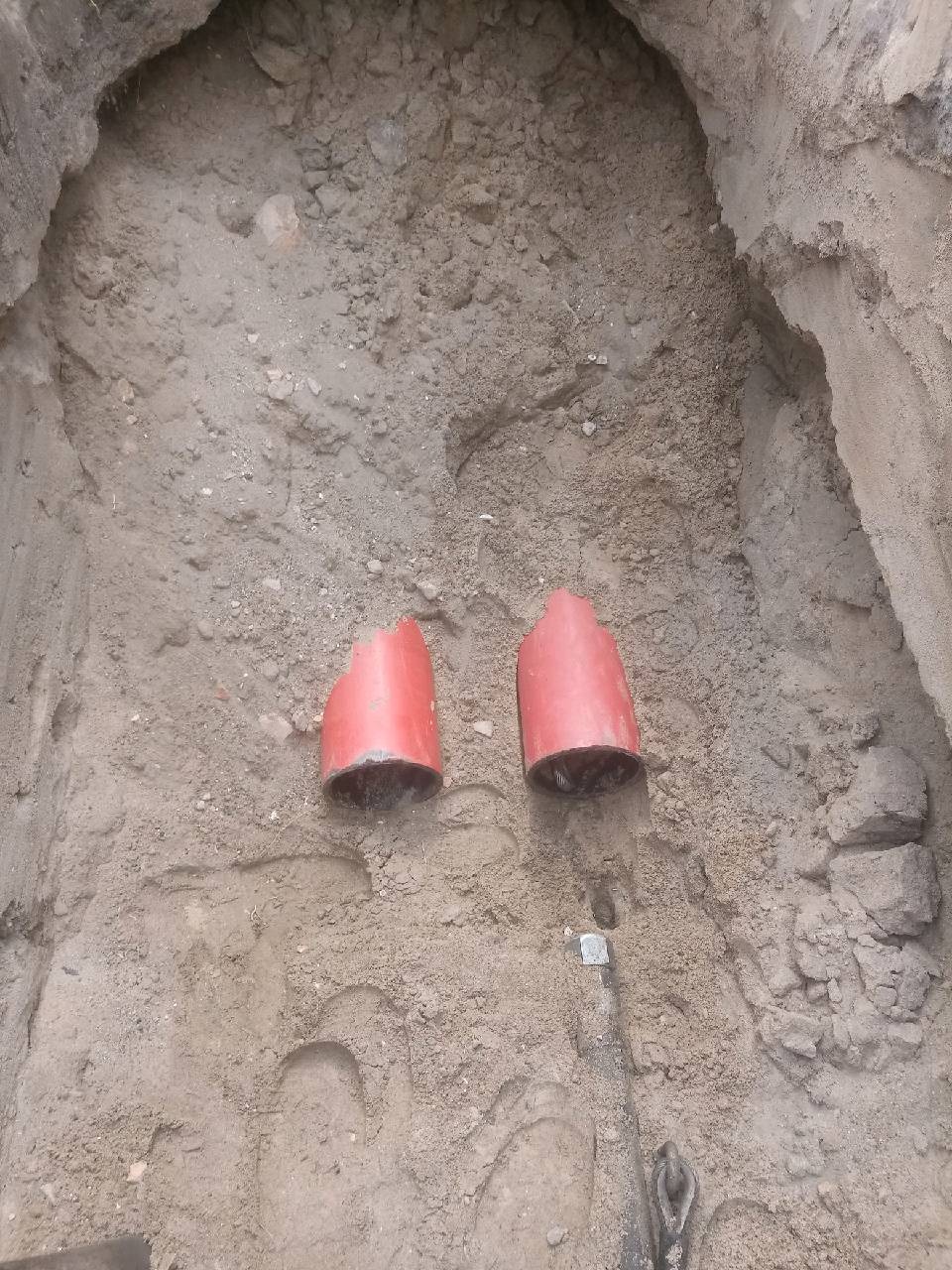Dwa czerwone kolanka rur w wykopie ziemnym, widoczny fragment rury i narzędzie, ziemia piaszczysta.