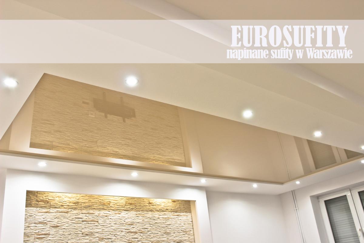 Błyszczący, beżowy sufit z wbudowanymi punktowymi lampami, z widoczną wnęką na ścianie wykończoną kamieniem dekoracyjnym w odcieniach beżu i brązu, oraz częściowo widocznym oknem.