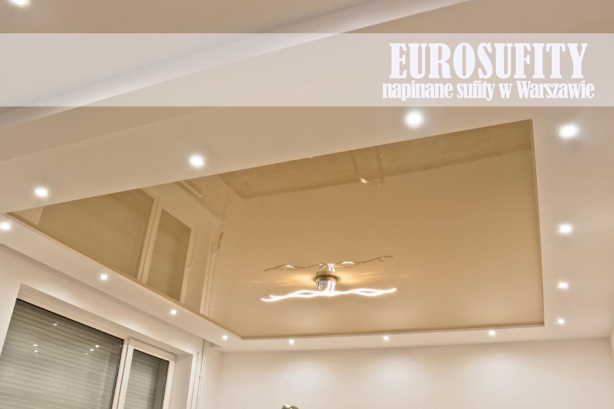 Beżowy, lustrzany sufit z wbudowanym oświetleniem punktowym i nowoczesną lampą w kształcie fali, widoczny z perspektywy pomieszczenia z oknem.