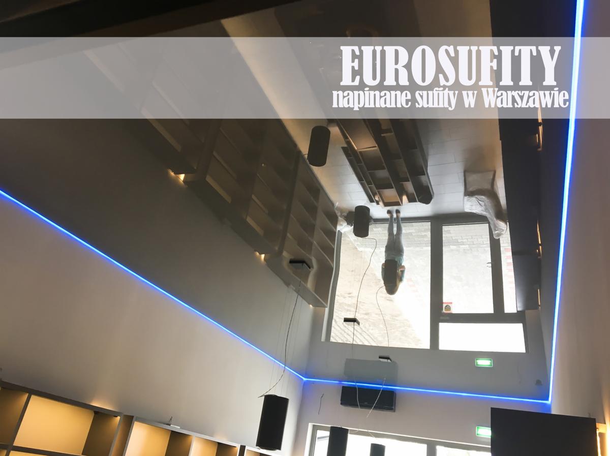 Błyszczący sufit z efektem lustra i niebieskim oświetleniem LED, odbijający wnętrze pomieszczenia z regałami i osobą stojącą przy oknie.