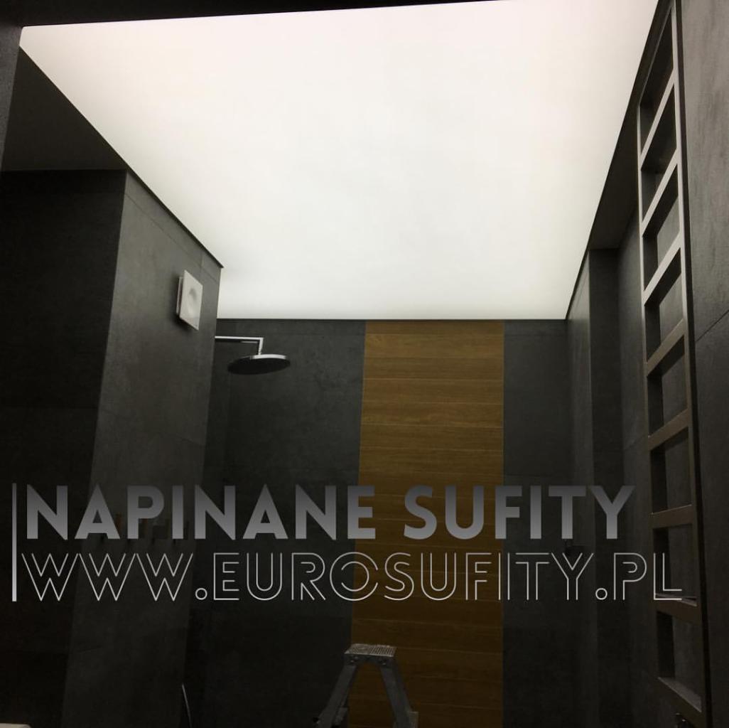 Nowoczesna łazienka z czarnymi płytkami i drewnianymi akcentami, oświetlona równomiernym, białym światłem z gładkiego sufitu. Widoczna drabinka dekoracyjna i minimalistyczna armatura.