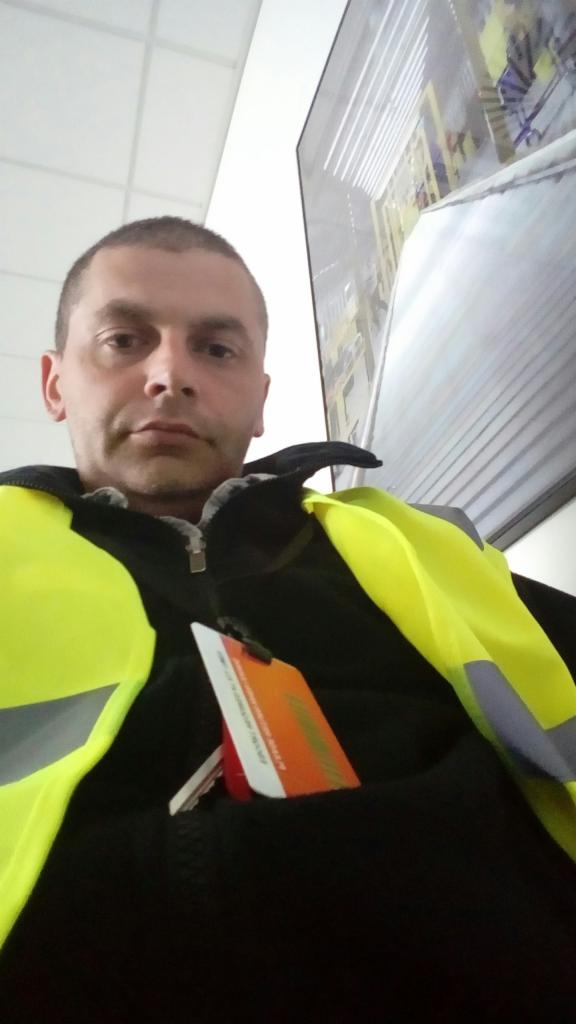 Mężczyzna w kamizelce odblaskowej z identyfikatorem w kieszeni, na tle zdjęcia magazynu z wózkami widłowymi.