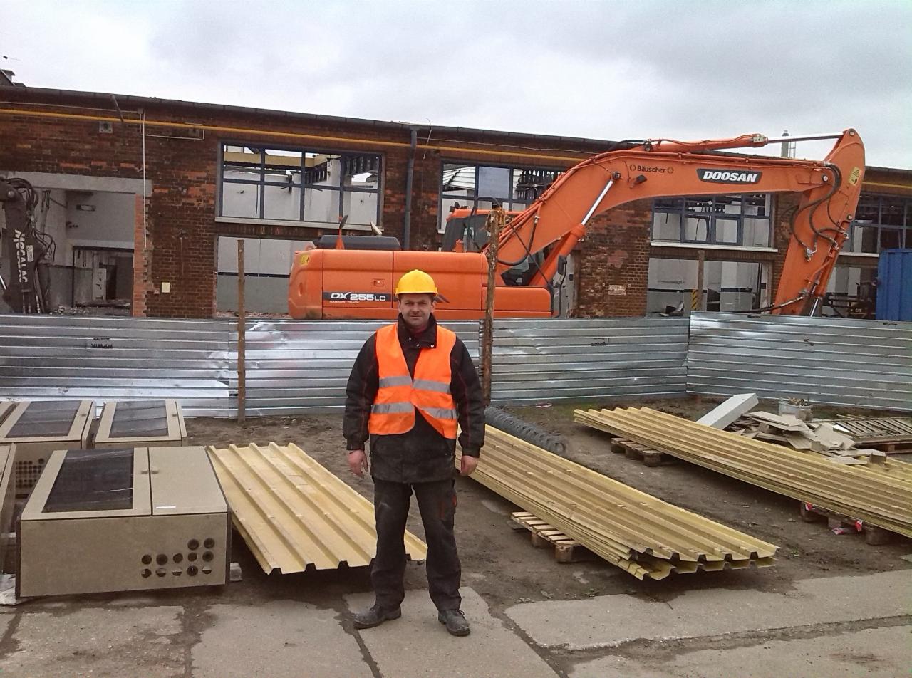 Pracownik w kamizelce odblaskowej i kasku na tle pomarańczowej koparki Doosan DX 255LC oraz budynku w trakcie rozbiórki, obok leżą płyty budowlane i metalowe panele.
