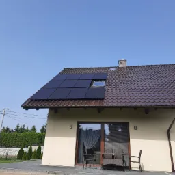 Dom jednorodzinny z brązową dachówką, na której zainstalowano czarne panele słoneczne. Widoczne okno dachowe i fragment tarasu z meblami ogrodowymi na tle błękitnego nieba.