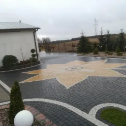 Ułożona kostka brukowa w kształcie kwiatu na podjeździe, obok budynku z ogrodem i elementami dekoracyjnymi z kostki.