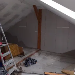 Poddasze w trakcie montażu płyt kartonowo-gipsowych, widoczne drewniane belki konstrukcyjne, drabina i narzędzia pracy.
