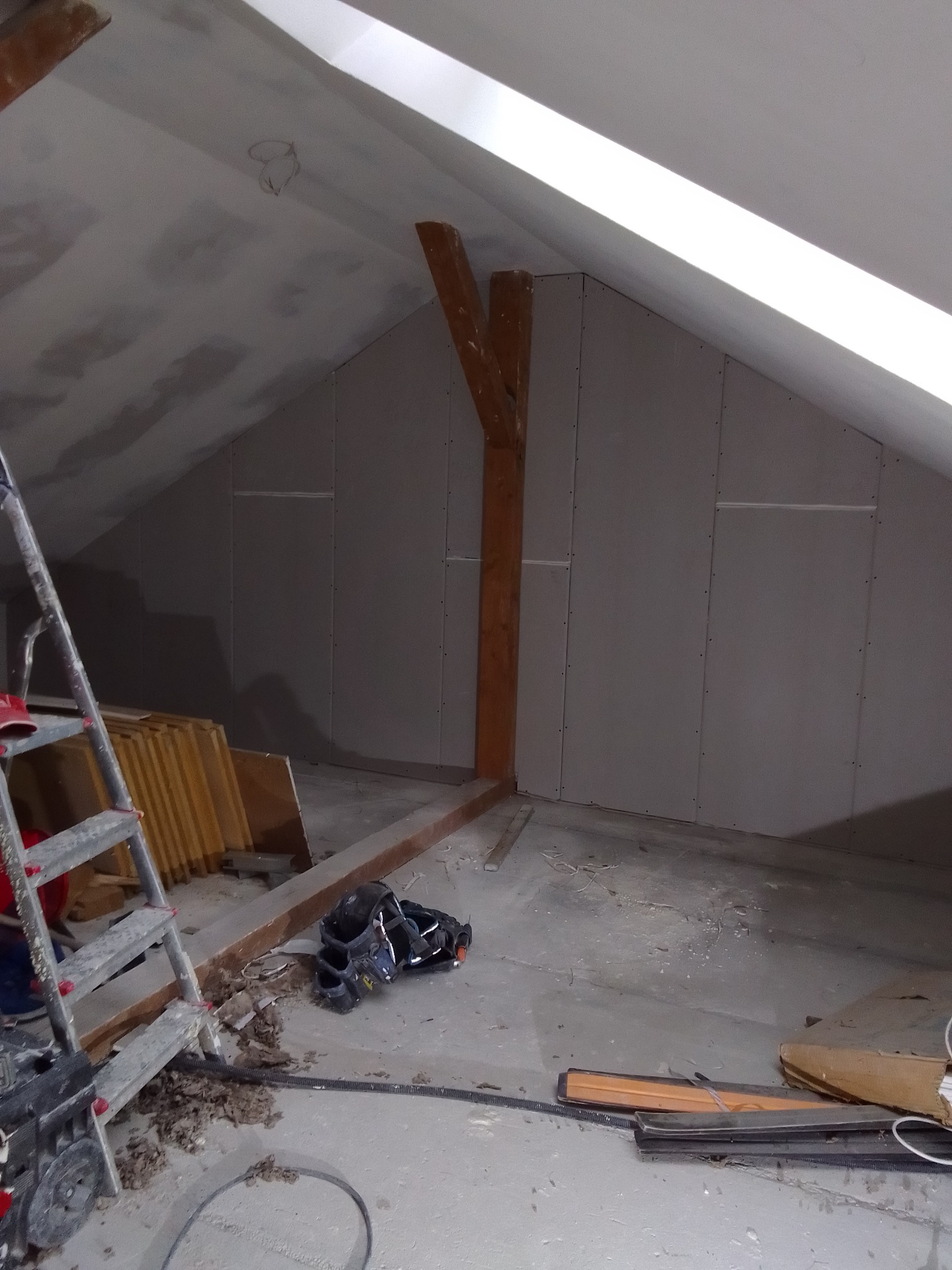 Poddasze w trakcie montażu płyt kartonowo-gipsowych, widoczne drewniane belki konstrukcyjne, drabina i narzędzia pracy.