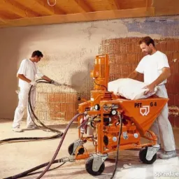 Tynkowanie maszynowe PFT G4 w trakcie aplikacji na ceglaną ścianę przez dwóch pracowników w białych uniformach. Widoczne węże i sufit z drewnianych belek.