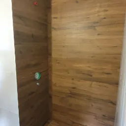 Prysznic w trakcie prac wykończeniowych, ściany i podłoga wyłożone płytkami imitującymi drewno, widoczne niedokończone przyłącza wodne.