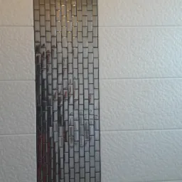 Ściana wyłożona białymi, poziomymi płytkami ceramicznymi z wstawką z mozaiki w odcieniach szarości, odbijającej światło.