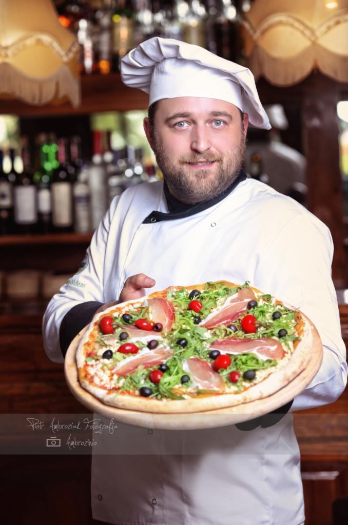 Uśmiechnięty kucharz w białym uniformie prezentuje pizzę na drewnianej desce, z szynką parmeńską, rukolą, czarnymi oliwkami i małymi czerwonymi papryczkami, na tle butelek z alkoholem i lampy.