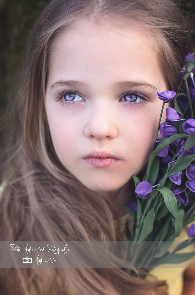 Portret dziewczynki o jasnych oczach trzymającej bukiet fioletowych kwiatów przy twarzy, z delikatnym rozmyciem tła.