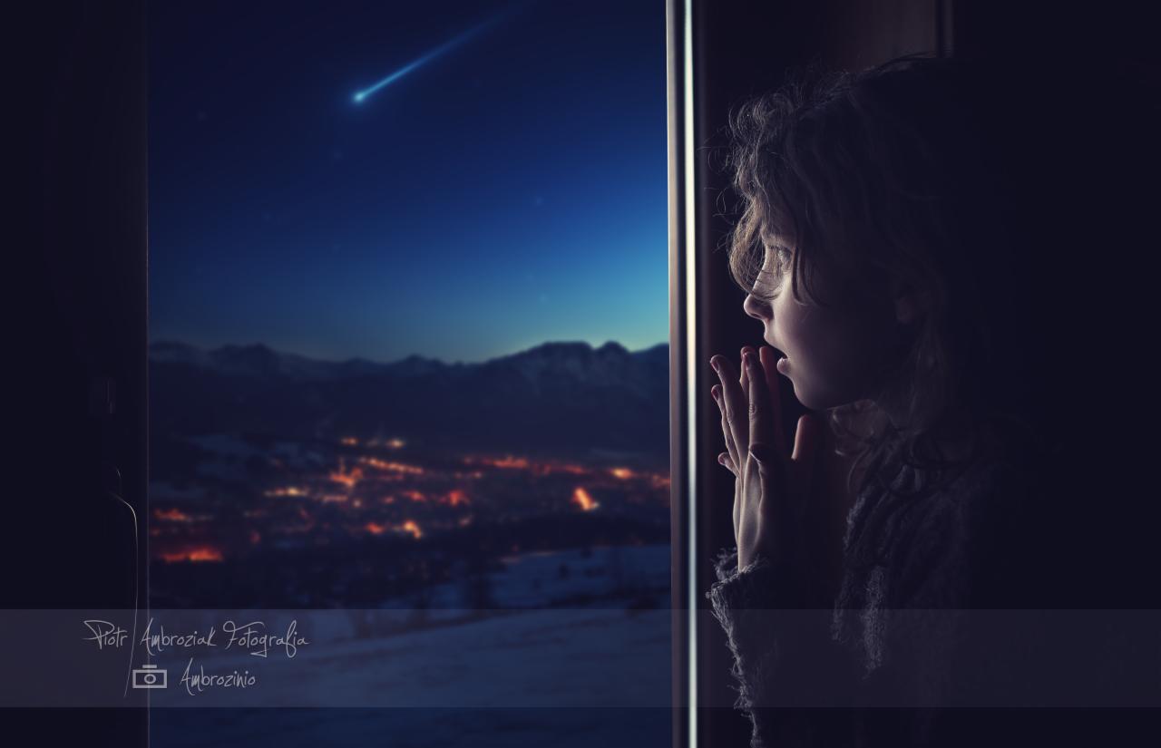 Dziewczynka z otwartymi ustami, przyciskająca dłonie do szyby okna, obserwuje spadającą gwiazdę nad nocnym miastem w górach.