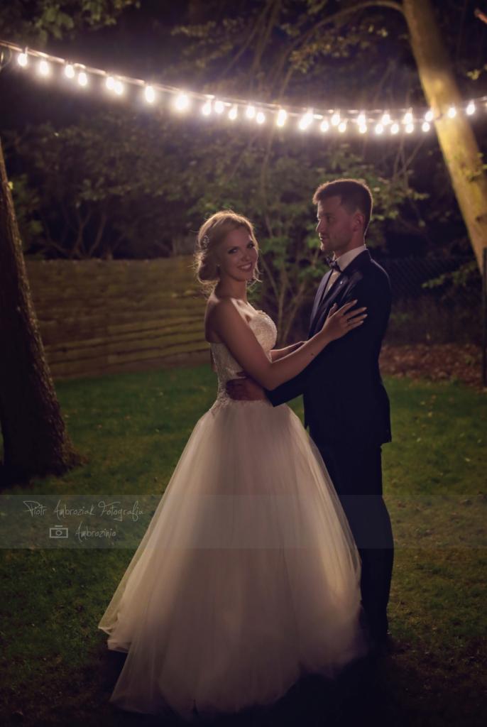 Para młoda w objęciach na tle drzew i girlandy światełek, panna młoda w sukni z koronkową górą i tiulowym dołem, pan młody w ciemnym garniturze i muszce.
