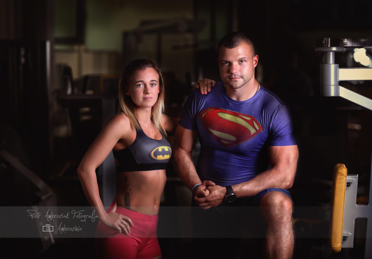 Para w strojach superbohaterów: kobieta w topie z logo Batmana, mężczyzna w koszulce Supermana, pozują w siłowni przy sprzęcie do ćwiczeń.