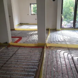 Rozłożona instalacja ogrzewania podłogowego w trakcie montażu, widoczne białe rury na folii z czerwoną siatką, zabezpieczone żółtą taśmą na obrzeżach pomieszczenia.