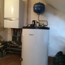 Kocioł gazowy Vaillant wiszący obok pionowego zbiornika na wodę, z widocznymi rurami miedzianymi i szarymi rurami kanalizacyjnymi, w pomieszczeniu z brązowymi płytkami.